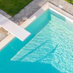 An aerial view of a rectangular swimming pool surrounded by a green lawn. A white diving board extends over the blue water.