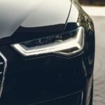 A close-up of a headlight on a black vehicle parked on a gray brick pathway. The background is blurry.