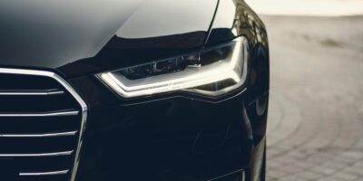 A close-up of a headlight on a black vehicle parked on a gray brick pathway. The background is blurry.