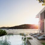 The sun sets on a modern, luxury beach house with a large rectangular pool. Sailboats float on the ocean in the background.