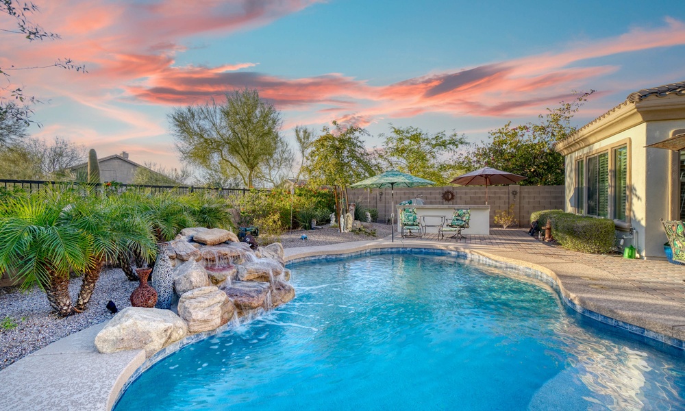 A circular backyard pool with a stone water feature surrounded by landscaping and two chairs and umbrellas.