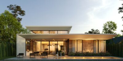 A long shot of a luxurious flat-roofed home with warm lighting illuminating its exterior walls and landscaping.