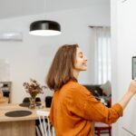 A smiling woman in an orange blouse presses the screen of a large touchscreen that hangs on the wall of a home.