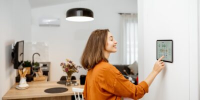 A smiling woman in an orange blouse presses the screen of a large touchscreen that hangs on the wall of a home.
