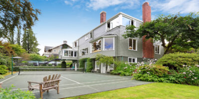 A modernly built house with a tennis court in the backyard. The backyard is beautifully landscaped with trees and plants.