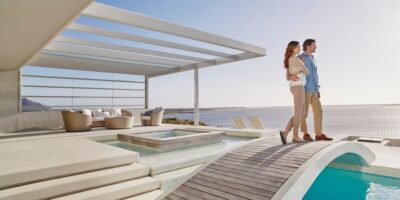 A young man and woman standing side by side on a bridge above a luxurious pool overlooking the ocean.