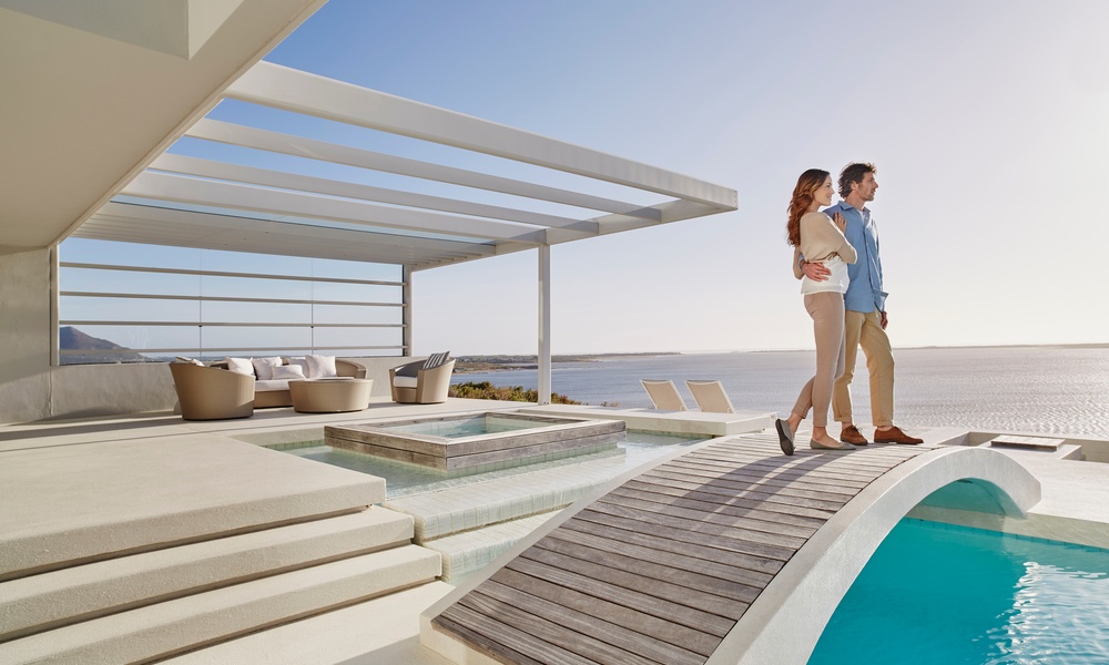 A young man and woman standing side by side on a bridge above a luxurious pool overlooking the ocean.
