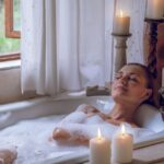 A woman closes her eyes as she enjoys a bubble bath. Around her are calming candles, plants, and an open window.