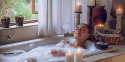 A woman closes her eyes as she enjoys a bubble bath. Around her are calming candles, plants, and an open window.