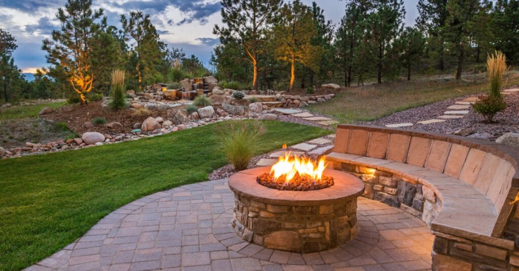 A fire bit is blazing with flames, there is a semi-circular bench stretching around the fire pit. There are trees in the background.