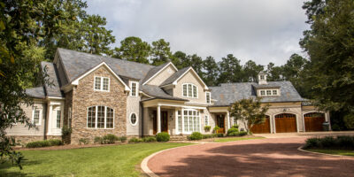 A brown gravel driveway leading up to a very large home, which features warm colors and a combination of brick and siding.