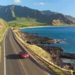 A convertible drives on a coastal road. The water on the right side is very blue, with mountains ahead of the car.