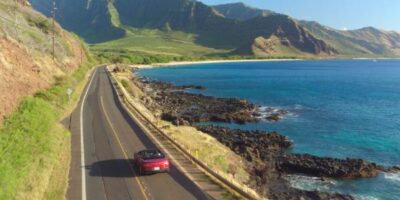 A convertible drives on a coastal road. The water on the right side is very blue, with mountains ahead of the car.