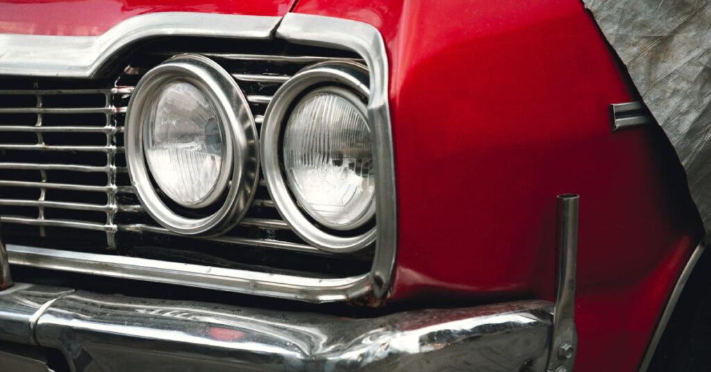 A close-up on the headlights of a classic, red car with silver detailing on the front grille and bumper.
