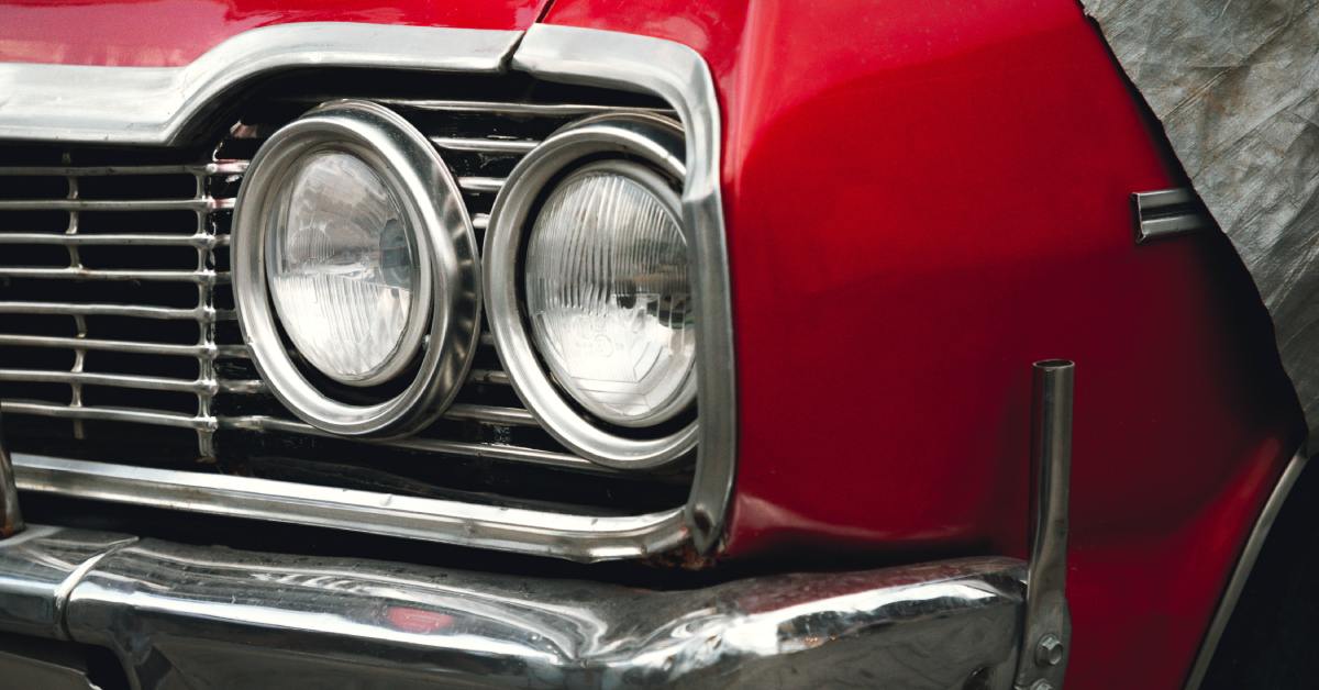 A close-up on the headlights of a classic, red car with silver detailing on the front grille and bumper.