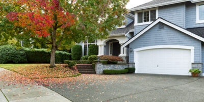 A blue house in autumn. There is a large tree next to the house with many fallen leaves surrounding it.