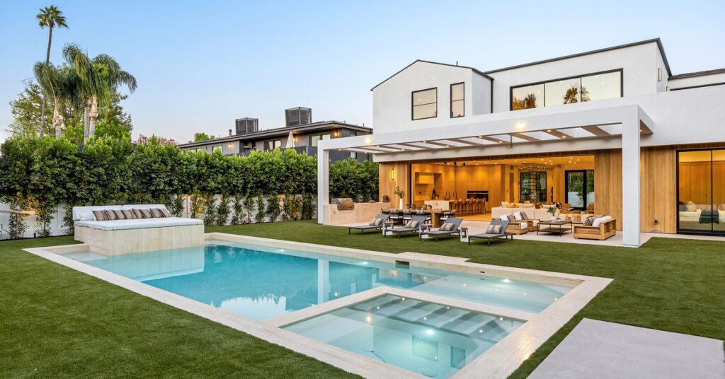 A view of a luxurious backyard with a resort-style pool, a hot tub, and an outdoor kitchen, surrounded by privacy trees.
