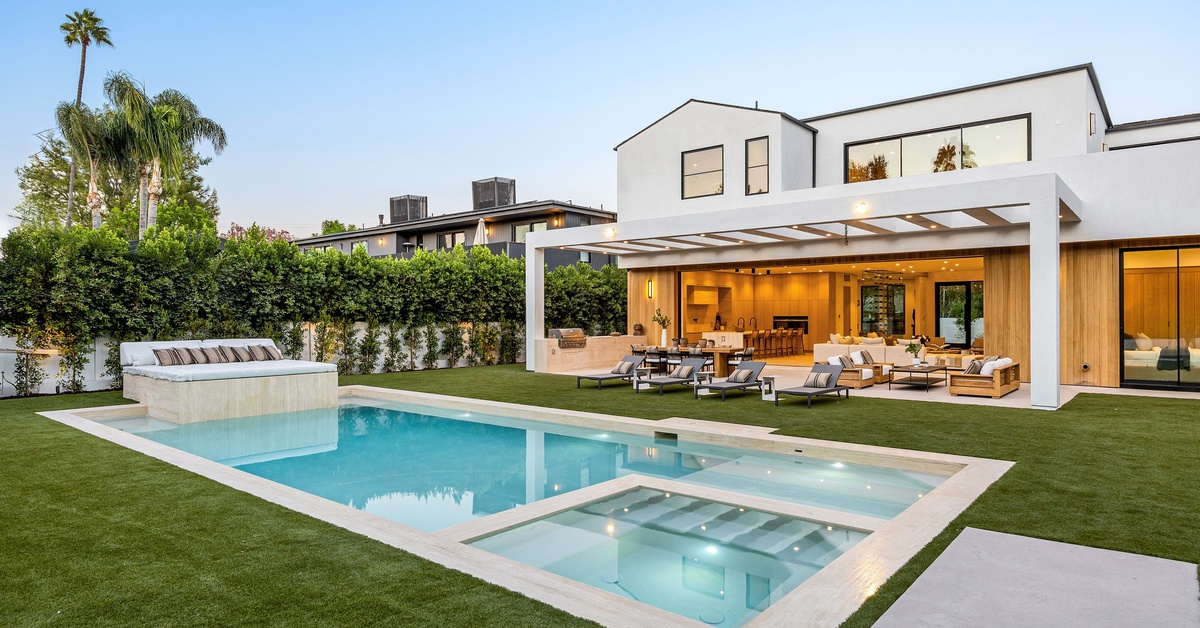 A view of a luxurious backyard with a resort-style pool, a hot tub, and an outdoor kitchen, surrounded by privacy trees.