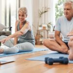 Two people stretching on yoga mats and looking at a computer on the floor. They are sitting on a wooden floor at home.