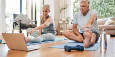 Two people stretching on yoga mats and looking at a computer on the floor. They are sitting on a wooden floor at home.