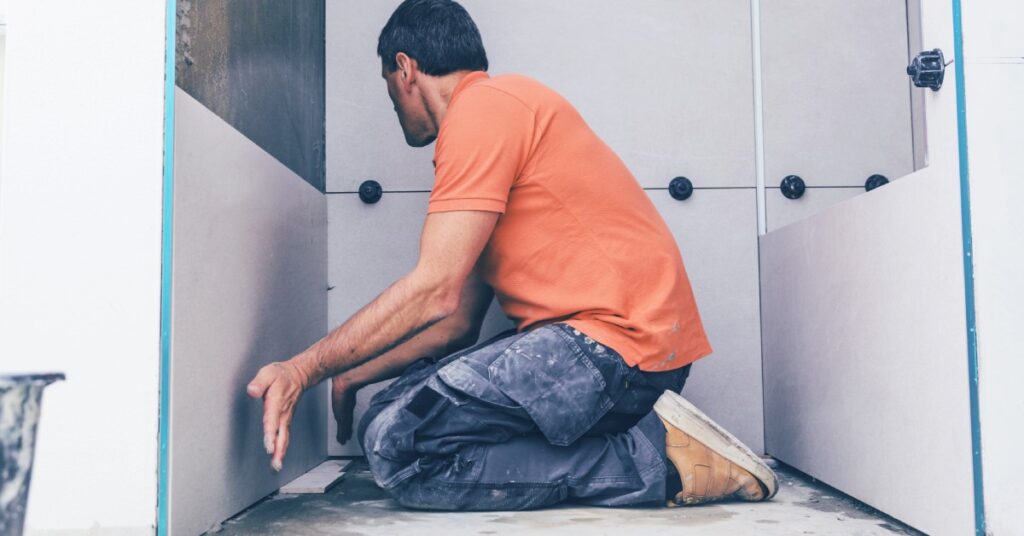 A man in an orange shirt works on assembling a bathroom panel, holding his hands close as he aligns the pieces.