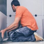 A man in an orange shirt works on assembling a bathroom panel, holding his hands close as he aligns the pieces.