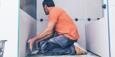 A man in an orange shirt works on assembling a bathroom panel, holding his hands close as he aligns the pieces.