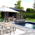 An elegantly landscaped backyard with a covered lounge area, swimming pool, fire feature, and a dining table on a tiled patio.