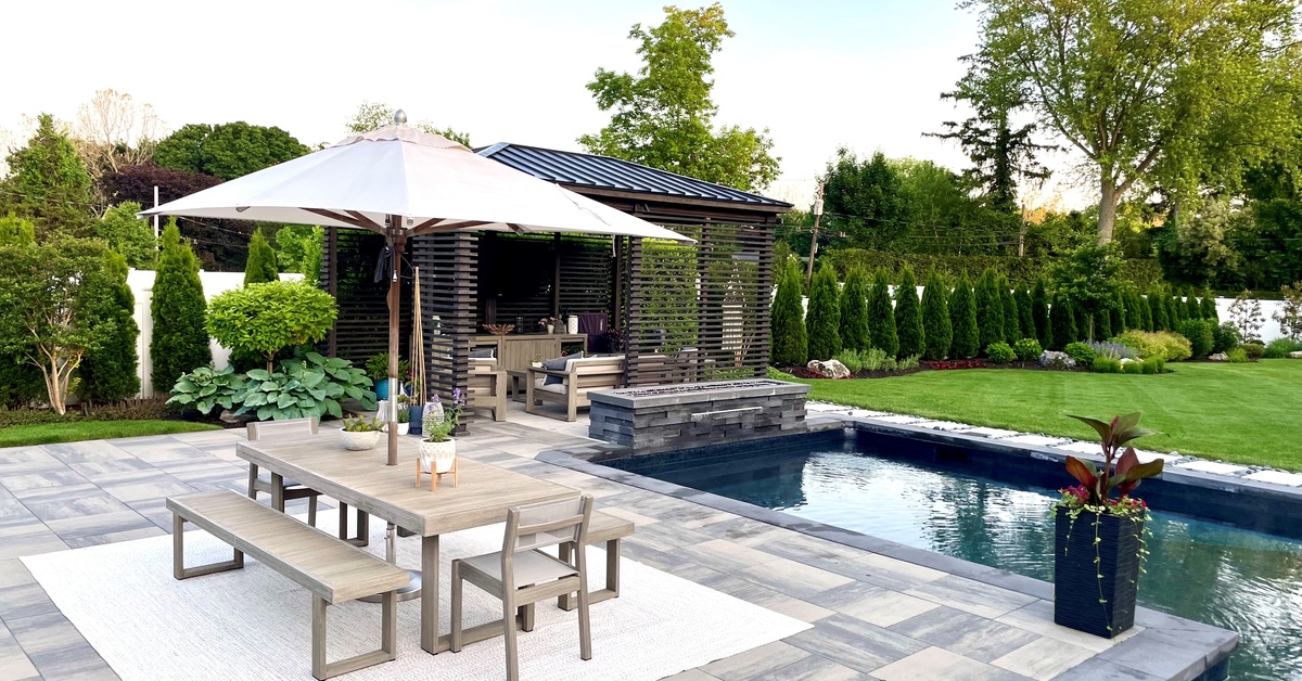 An elegantly landscaped backyard with a covered lounge area, swimming pool, fire feature, and a dining table on a tiled patio.