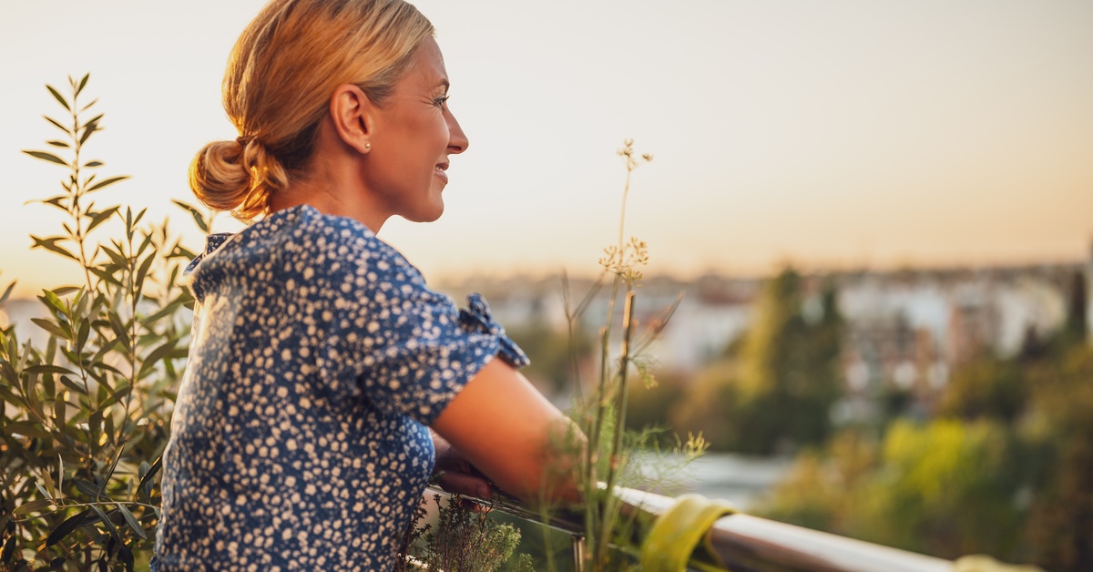 A woman wearing a blue dress stands on a balcony with plants, overlooking a blurred city view at golden hour.