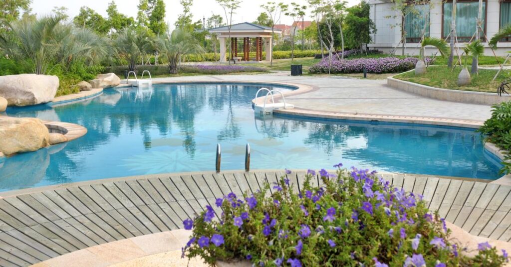 A curved backyard pool with clear blue water, purple flowers in the foreground, lush landscaping, and a gazebo.