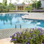 A curved backyard pool with clear blue water, purple flowers in the foreground, lush landscaping, and a gazebo.
