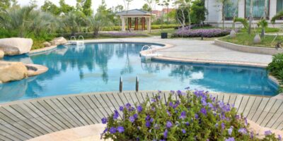 A curved backyard pool with clear blue water, purple flowers in the foreground, lush landscaping, and a gazebo.