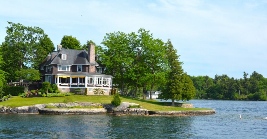 A large lakeside house on a manicured green lawn lined with trees, overlooking calm blue water.