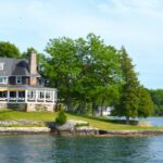 A large lakeside house on a manicured green lawn lined with trees, overlooking calm blue water.