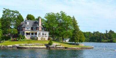 A large lakeside house on a manicured green lawn lined with trees, overlooking calm blue water.