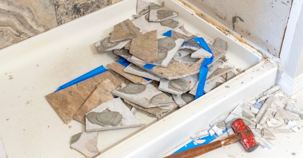 A tiled shower with the tiles on one of the walls smashed and sitting in the tub during demolition in a bathroom.