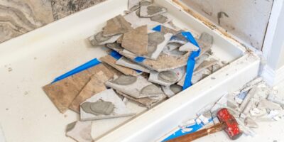 A tiled shower with the tiles on one of the walls smashed and sitting in the tub during demolition in a bathroom.