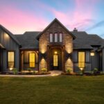 A modern luxury home features a stone entry, dark siding, large windows, and warm exterior lighting at sunset.