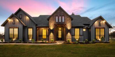 A modern luxury home features a stone entry, dark siding, large windows, and warm exterior lighting at sunset.