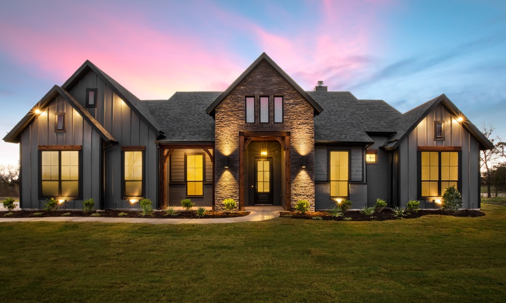 A modern luxury home features a stone entry, dark siding, large windows, and warm exterior lighting at sunset.