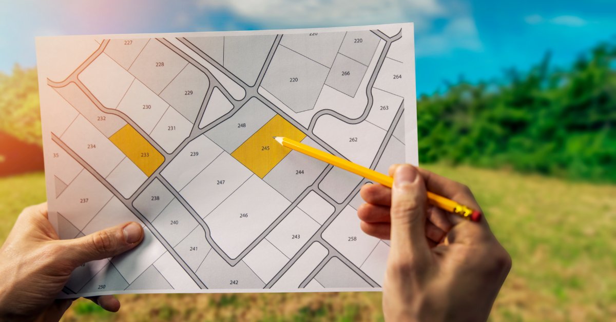 Two hands holding up a land blueprint and using a pencil to point out a specific plot of land. A field is right behind them.