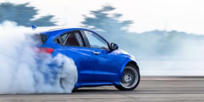 A blue car drifts on a race track, producing lots of smoke from its rear tires. Trees can be seen in the background.
