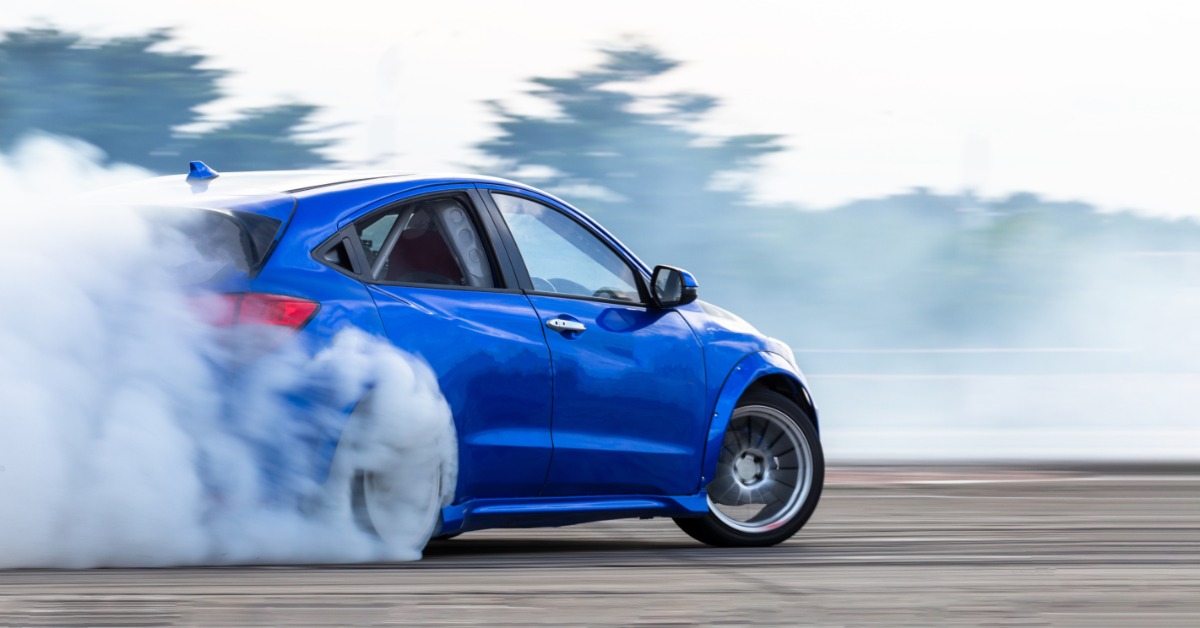 A blue car drifts on a race track, producing lots of smoke from its rear tires. Trees can be seen in the background.