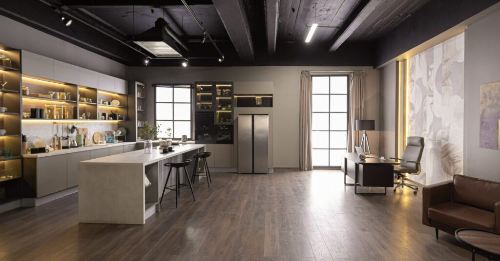 A modern kitchen and office share an open luxury room with dark ceilings, wood floors, and bright display cabinets.