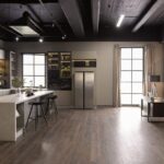 A modern kitchen and office share an open luxury room with dark ceilings, wood floors, and bright display cabinets.