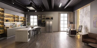 A modern kitchen and office share an open luxury room with dark ceilings, wood floors, and bright display cabinets.