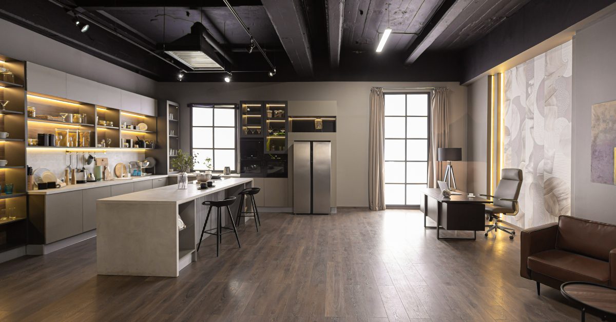 A modern kitchen and office share an open luxury room with dark ceilings, wood floors, and bright display cabinets.