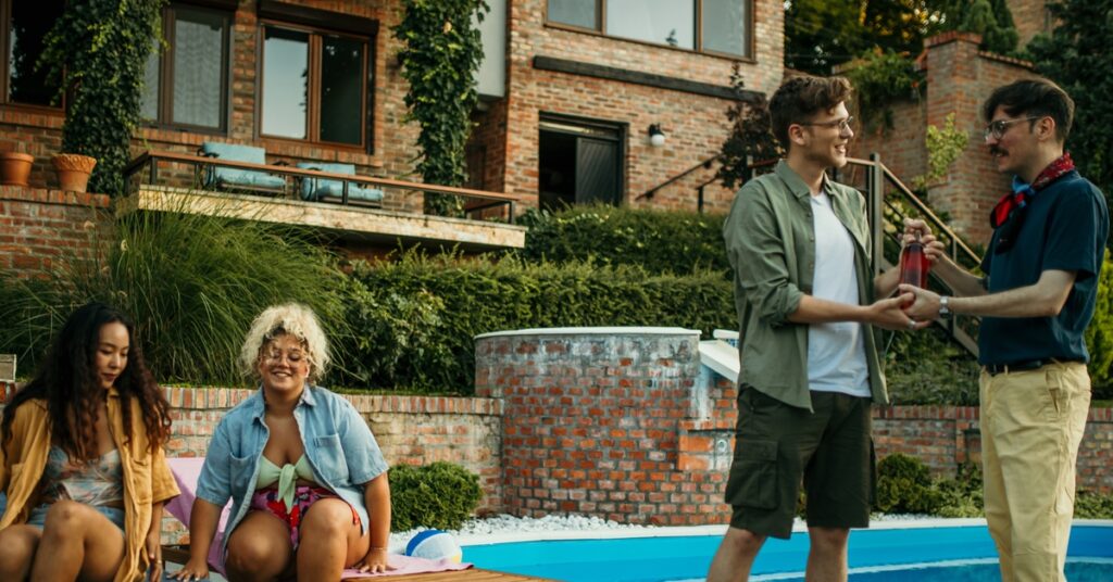 a group of adults talking and holding drinks near a swimming pool during an outdoor backyard gathering.