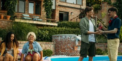a group of adults talking and holding drinks near a swimming pool during an outdoor backyard gathering.